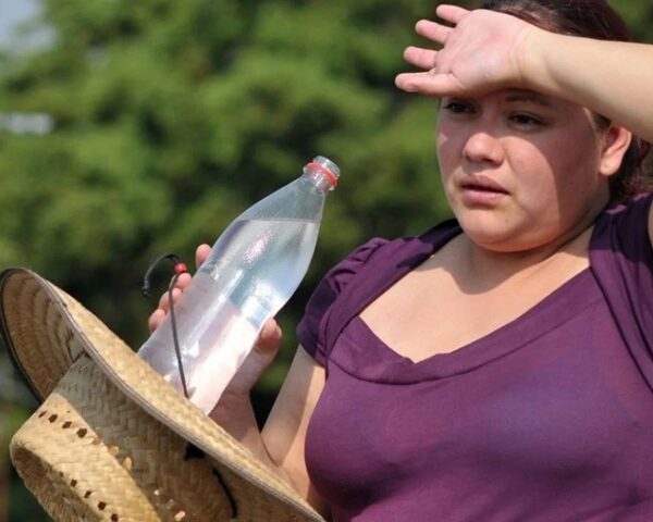 Woman shielding her eyes from bright sun while resting outdoors, holding a plastic water bottle and a straw hat on her knee.