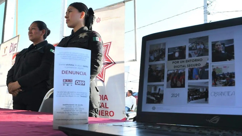 Two female police officers stand at a community safety event; a laptop screen shows a digital-safety collage, and a brochure labeled 'DENUNCIA 089' rests on the table.