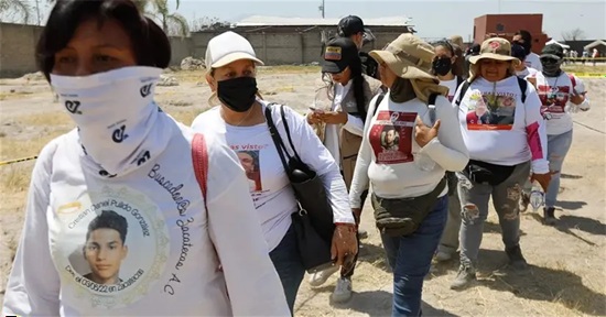 Group of people walking in a dusty outdoor area, wearing white shirts with a portrait and some masks or face coverings, as part of a march or vigil.