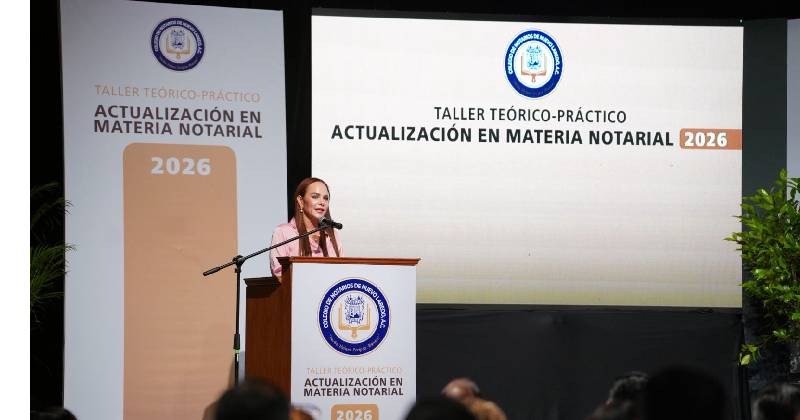 Female speaker at a podium giving a talk at a theoretical-practical workshop on notarial matters (2026), with banners displaying 'Actualización en Materia Notarial' and official emblems.