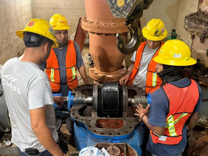 Four workers in hard hats and safety vests assemble a large blue valve on a metal pipe system.