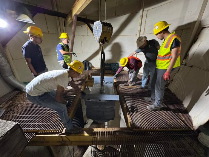 Construction crew in hard hats guides a heavy metal block using a crane in an industrial room in lift/setup work.