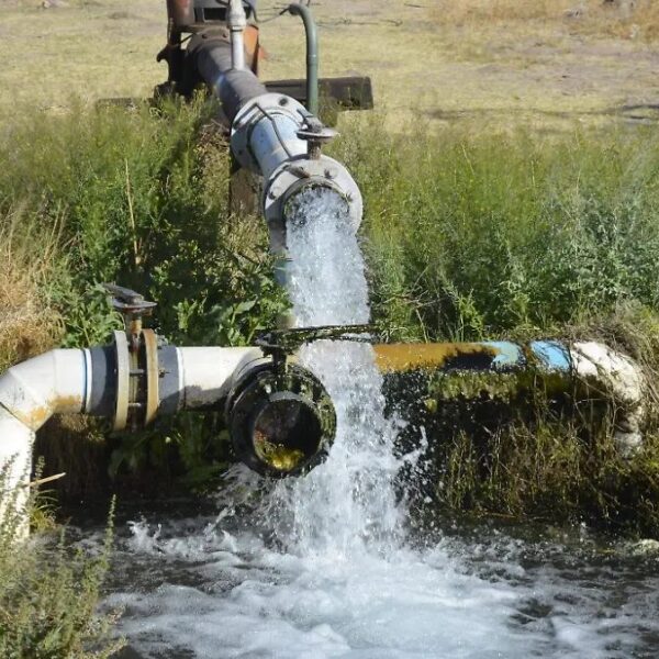 Water jet spraying from an industrial pipe into a small ditch, with a valve and metal fittings nearby in a grassy area.] ,