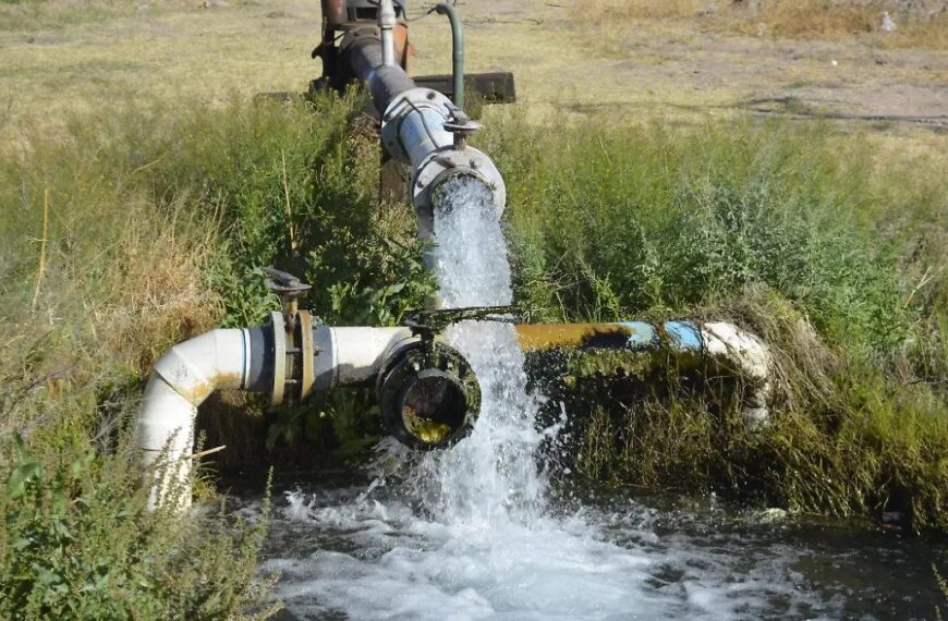 Water jet spraying from an industrial pipe into a small ditch, with a valve and metal fittings nearby in a grassy area.] ,