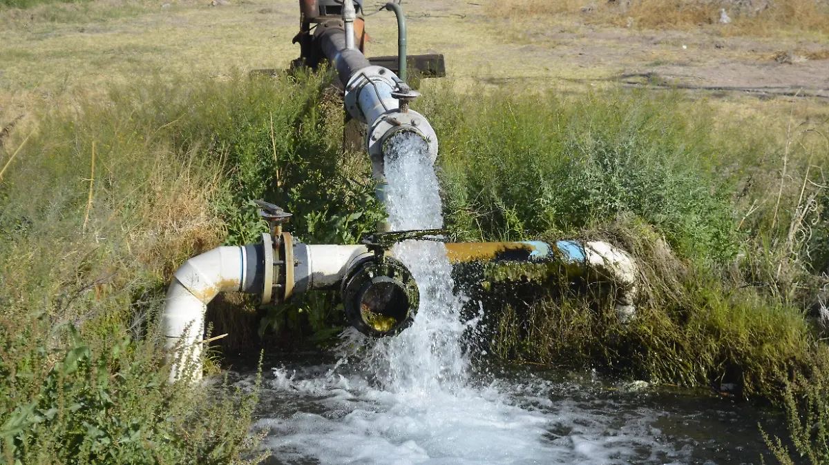 Water jet spraying from an industrial pipe into a small ditch, with a valve and metal fittings nearby in a grassy area.] ,