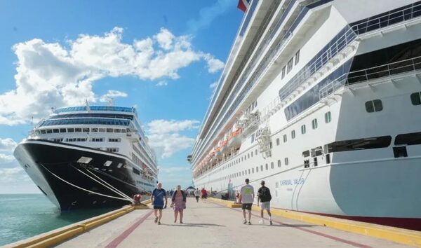 Two large cruise ships docked at a sunny port while passengers walk along the pier between them.