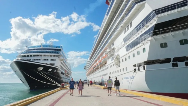 Two large cruise ships docked at a sunny port while passengers walk along the pier between them.