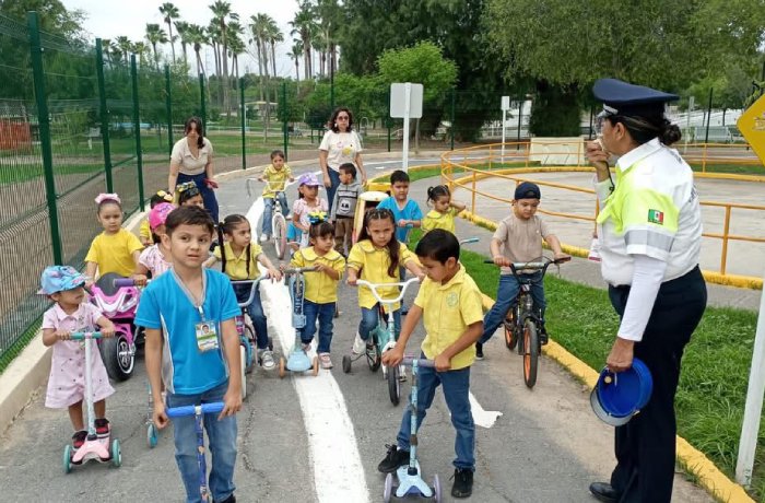 Group of children learning bicycle and scooter safety with a police officer supervising on a closed road path outside a park.
