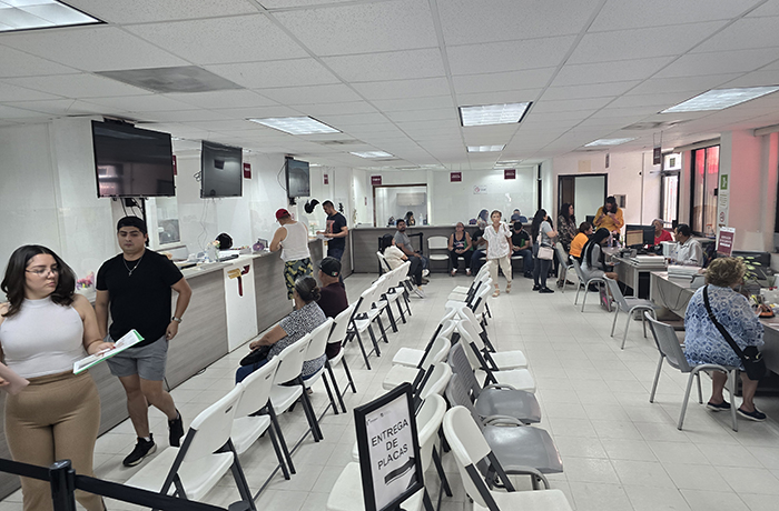 Busy service center with people waiting in white chairs and staff at desks; sign reads 'ENTREGA DE PLACAS'.