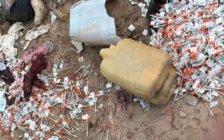 Outdoor ground littered with discarded syringes and two large plastic jugs on dusty earth.