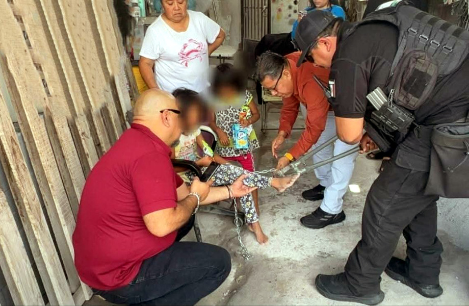 Authorities restrain a young person on the ground with metal restraints as bystanders watch in a market-alley setting (chains around wrists/legs visible).