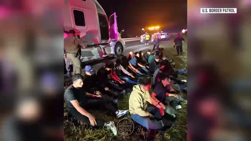 Group of people seated on grass at night beside a stopped truck, with border patrol in the background (U.S. Border Patrol).
