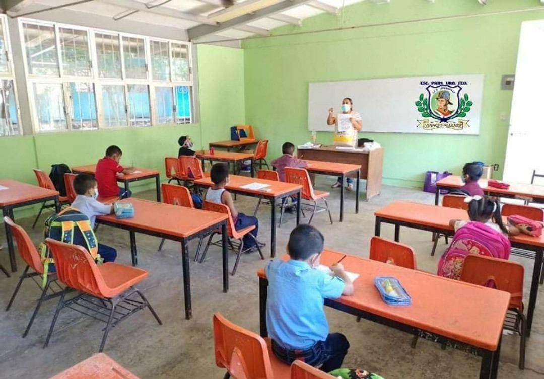 Teacher wearing a mask instructing elementary students seated at orange desks in a bright green classroom.