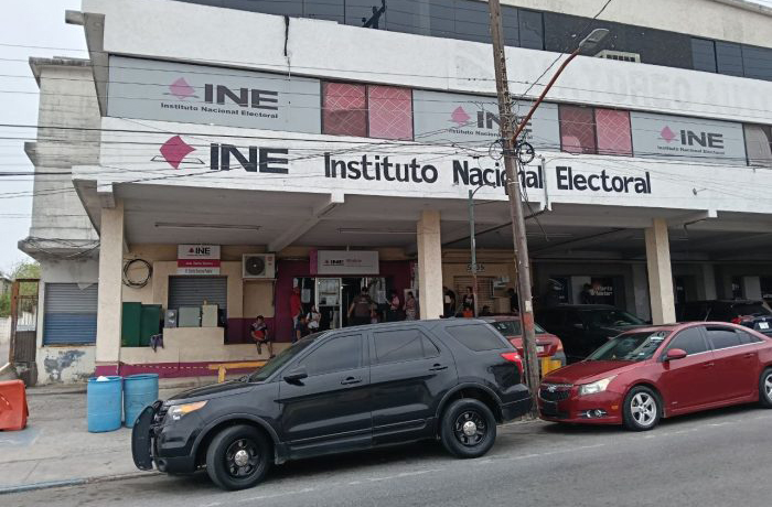 Front view of an Instituto Nacional Electoral (INE) building with large signs, people at the entrance, and a black SUV and a red car parked on the street.