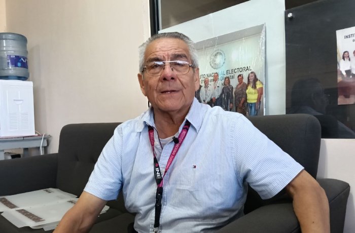 Older man in a light blue short-sleeve shirt sits on a dark sofa in an office, wearing a lanyard with a badge around his neck.
