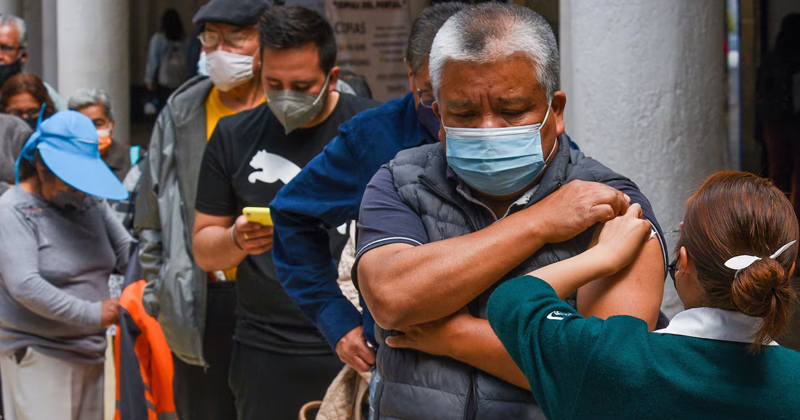 Man in a mask receives a vaccination from a clinician while others in line wear masks behind him.