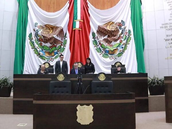 Legislative chamber with five officials seated at a dark wooden dais, two large Mexican flags behind them.