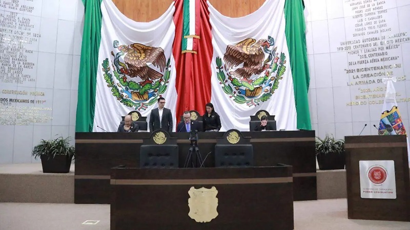 Legislative chamber with five officials seated at a dark wooden dais, two large Mexican flags behind them.