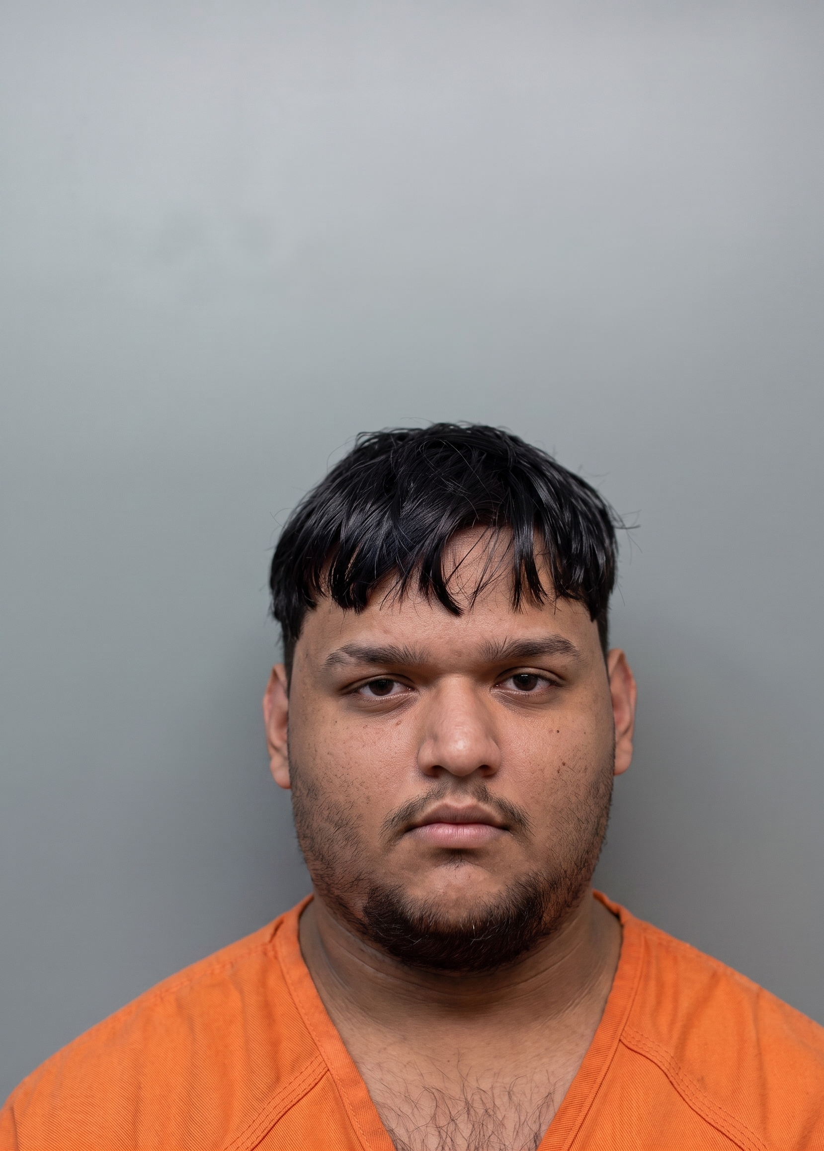 Portrait of a young man with dark hair and stubble wearing an orange jail jumpsuit against a gray background.