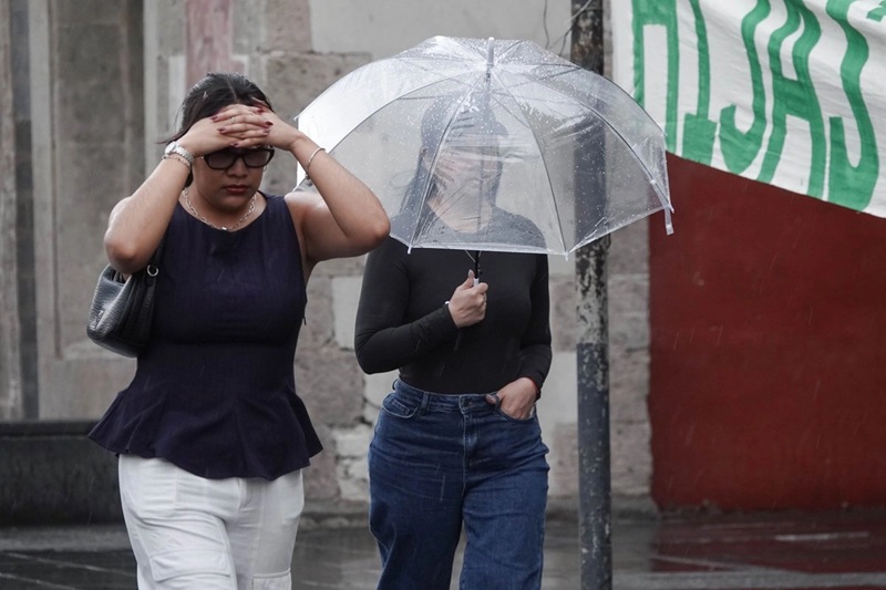 Two women walk in the rain; one shields her face with her hands while the other holds a clear umbrella on a wet street.