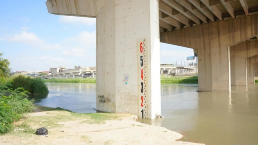 Bridge pillar with a vertical height gauge (numbers 1–6) beside a calm river under an overpass, clear sky above, sandy bank nearby.