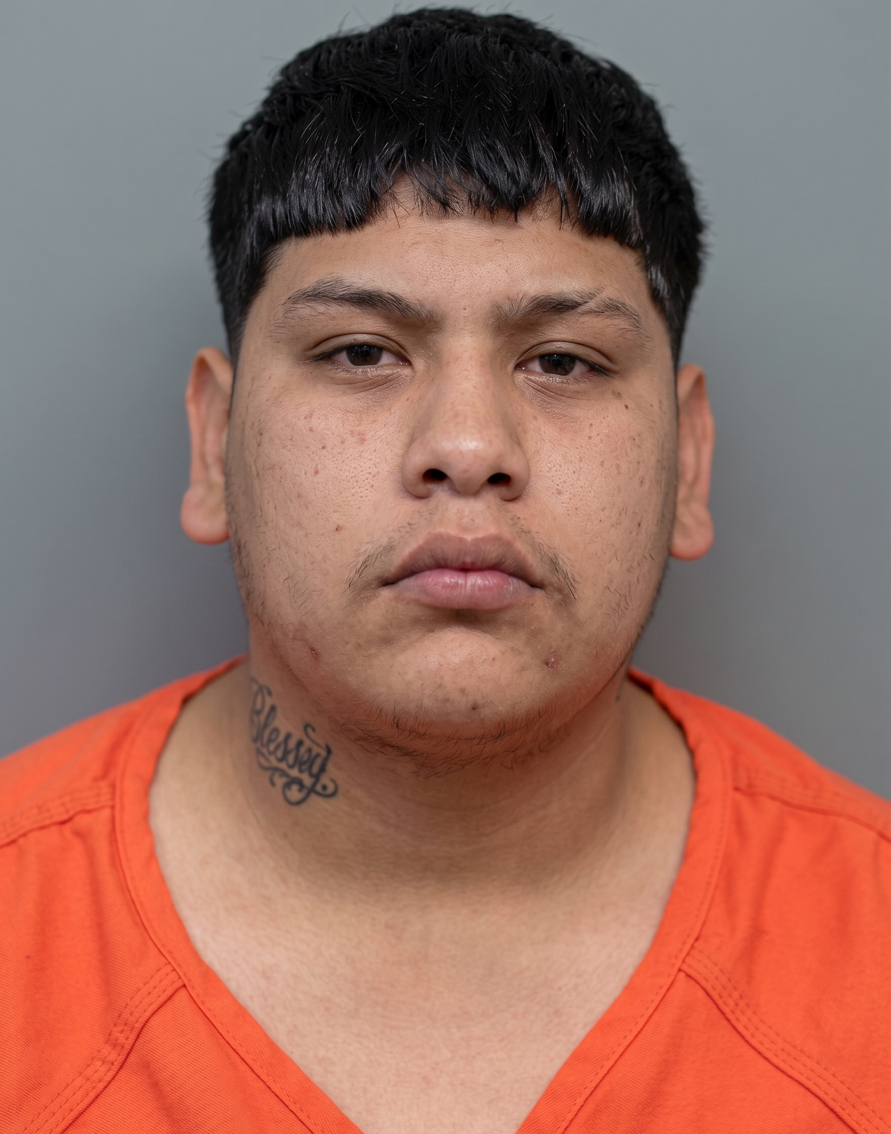 Mugshot of a young man with short black hair wearing an orange jumpsuit, facing the camera with a neutral expression, gray background, tattoo on neck reading 'Blessed'