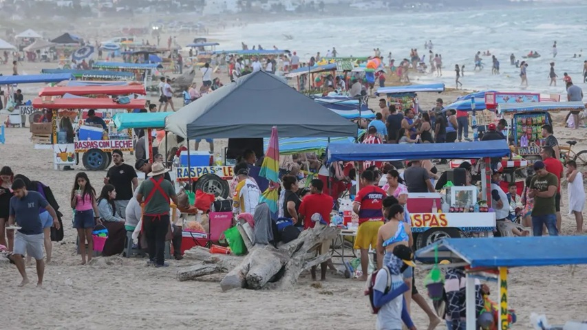 Crowded beach scene with colorful vendor stalls under canopies, selling snacks; families and vendors on sandy shore with the sea in the background. Signs advertise raspas (snack stands).
