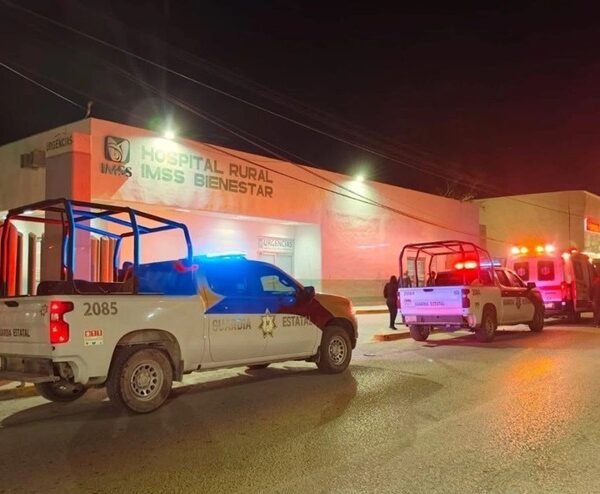 Nighttime scene at a rural hospital with emergency vehicles parked outside and blue and red lights flashing in front of the building labeled 'Hospital Rural IMSS Bienestar'.