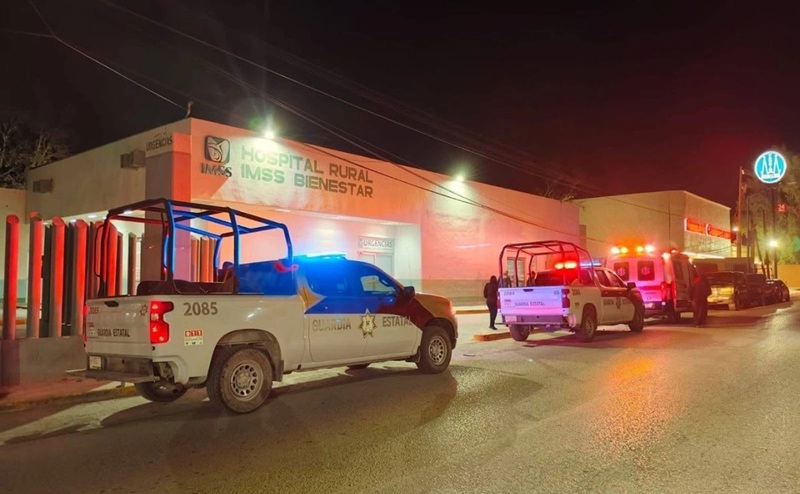Nighttime scene at a rural hospital with emergency vehicles parked outside and blue and red lights flashing in front of the building labeled 'Hospital Rural IMSS Bienestar'.