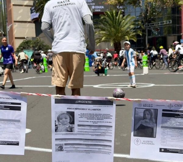 Man in a gray shirt and beige shorts, seen from behind, standing at a city street event with posters on a barrier in the foreground.