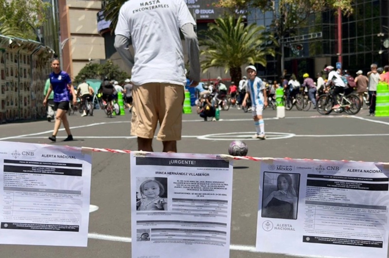 Man in a gray shirt and beige shorts, seen from behind, standing at a city street event with posters on a barrier in the foreground.