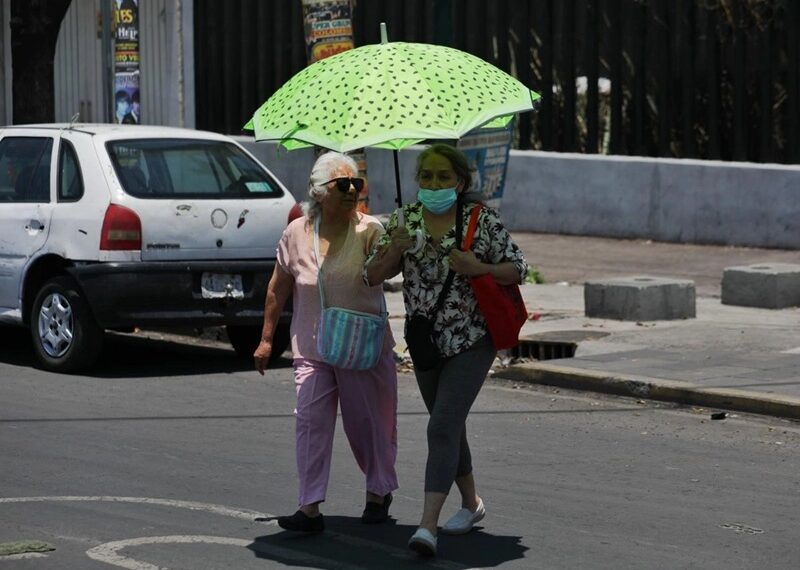 Two women walk side by side in the street under a bright green umbrella; one wears pink and carries a striped bag, the other wears a mask and a floral top with a red bag.