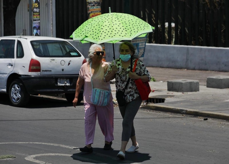 Two women walk side by side in the street under a bright green umbrella; one wears pink and carries a striped bag, the other wears a mask and a floral top with a red bag.