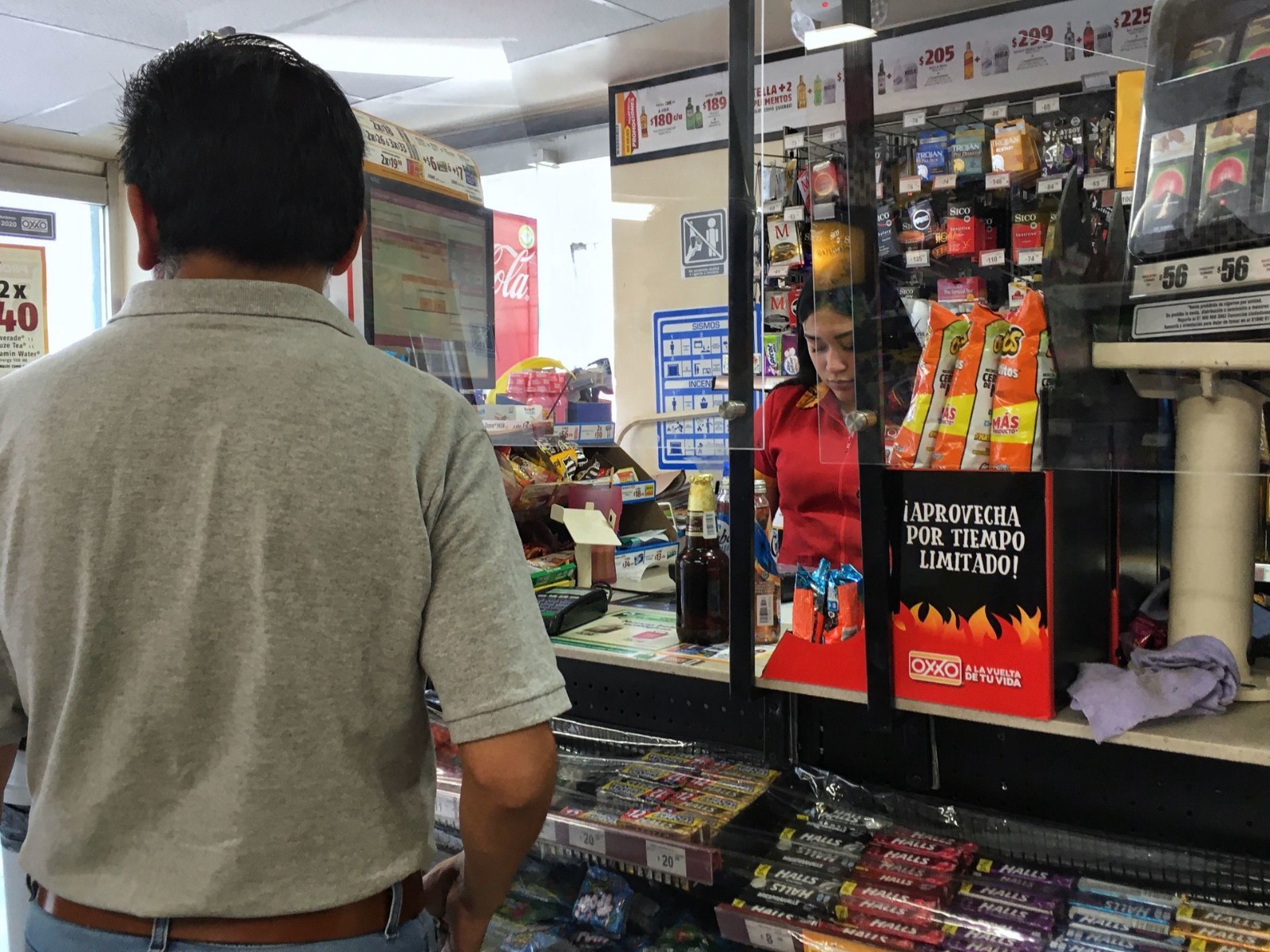 Customer waits at a convenience store register, cashier behind a glass barrier sorting items, shelves of snacks and drinks in view.