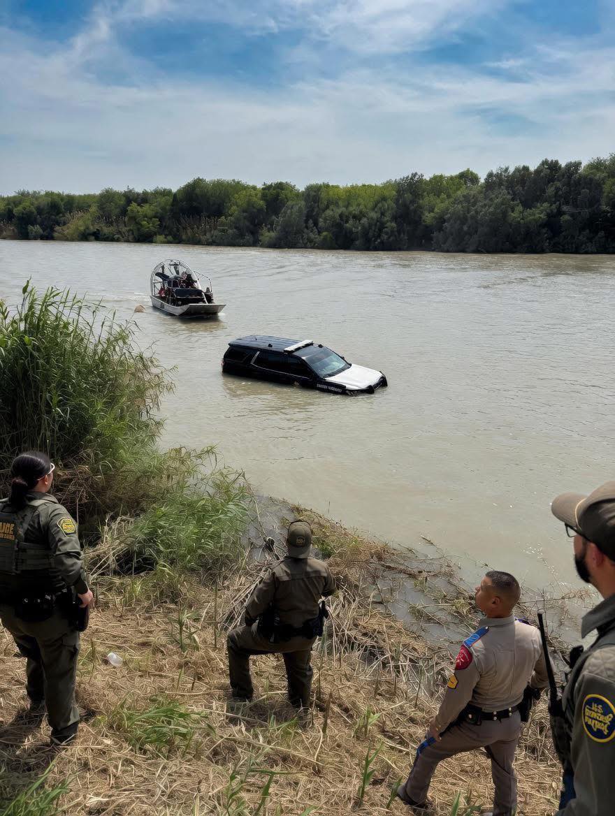 Police and rescue personnel stand on a riverbank overlooking a partially submerged black and white SUV in the water while a small boat approaches the scene.