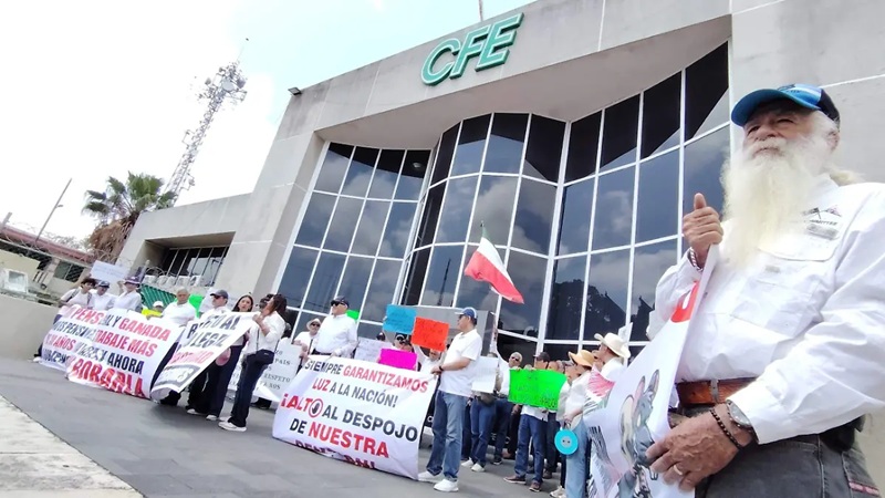 Group of protesters outside a CFE building, holding banners; a bearded man in the foreground gives a thumbs up.