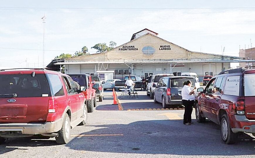 Border crossing plaza in front of Aduana Frontera Nuevo Laredo with cars parked in a lot and a few people near a red truck.
