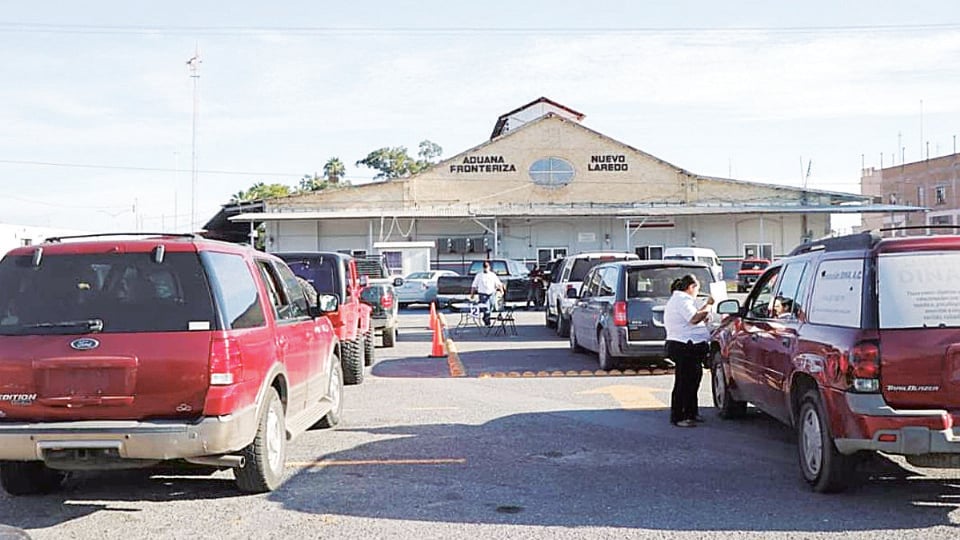 Border crossing plaza in front of Aduana Frontera Nuevo Laredo with cars parked in a lot and a few people near a red truck.