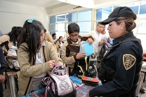 Police officer in uniform oversees students inspecting backpacks at a school safety event in a classroom setting.