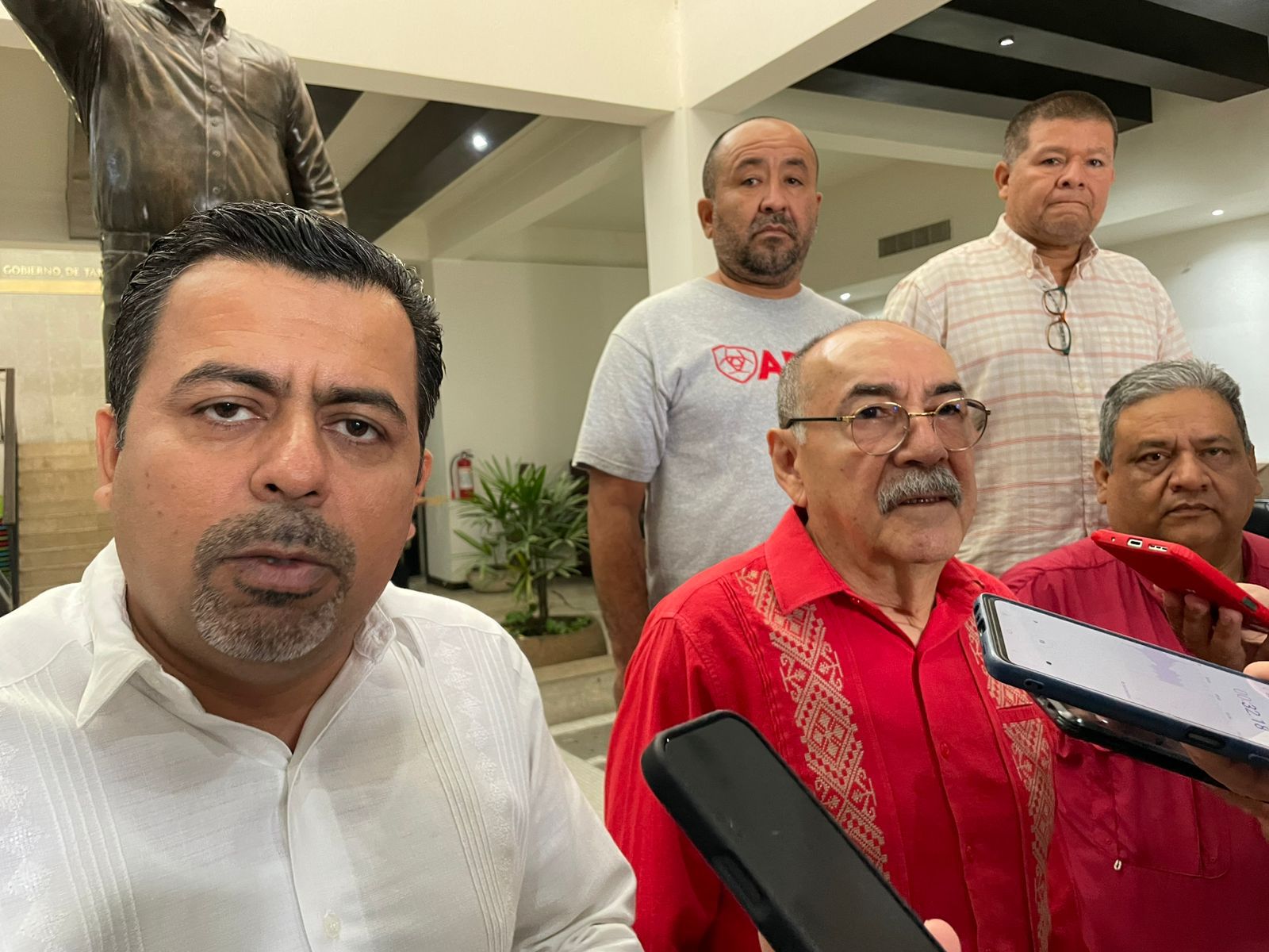 Group of five men indoors; a man in a white shirt takes a selfie while reporters hold phones and a microphone nearby in a lobby setting.