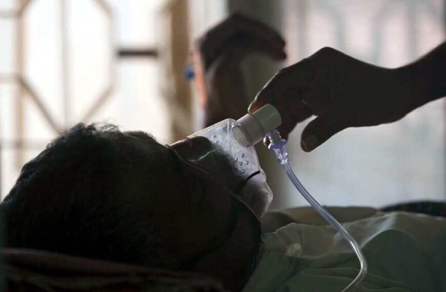 Patient lying in bed with an oxygen mask connected to tubing, as a gloved hand adjusts the mask.