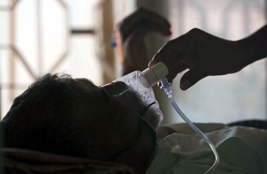 Patient lying in bed with an oxygen mask connected to tubing, as a gloved hand adjusts the mask.
