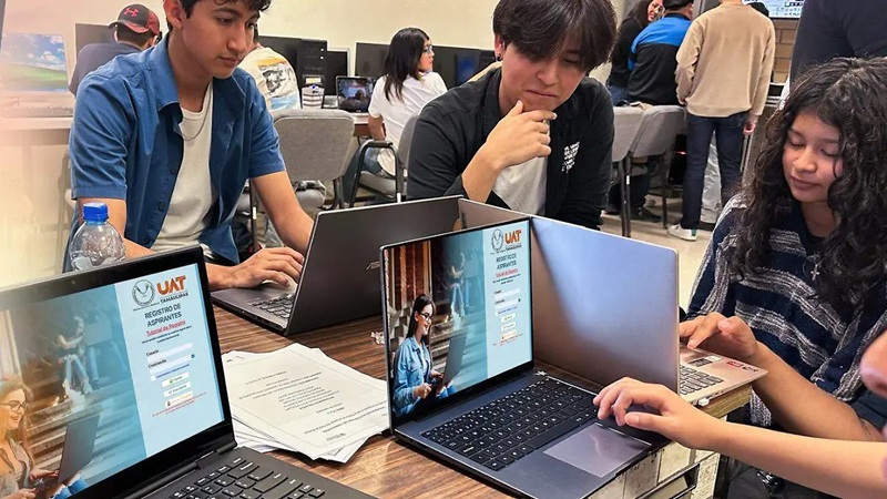Group of students gathered around a table in a computer lab, laptops open and screens visible as they work together on a project.