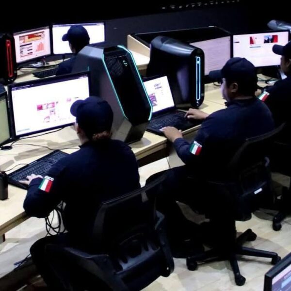 Group of operators in dark uniforms sit at a row of desks with monitors and illuminated PC towers in a computer lab, Italian flag patches on sleeves.