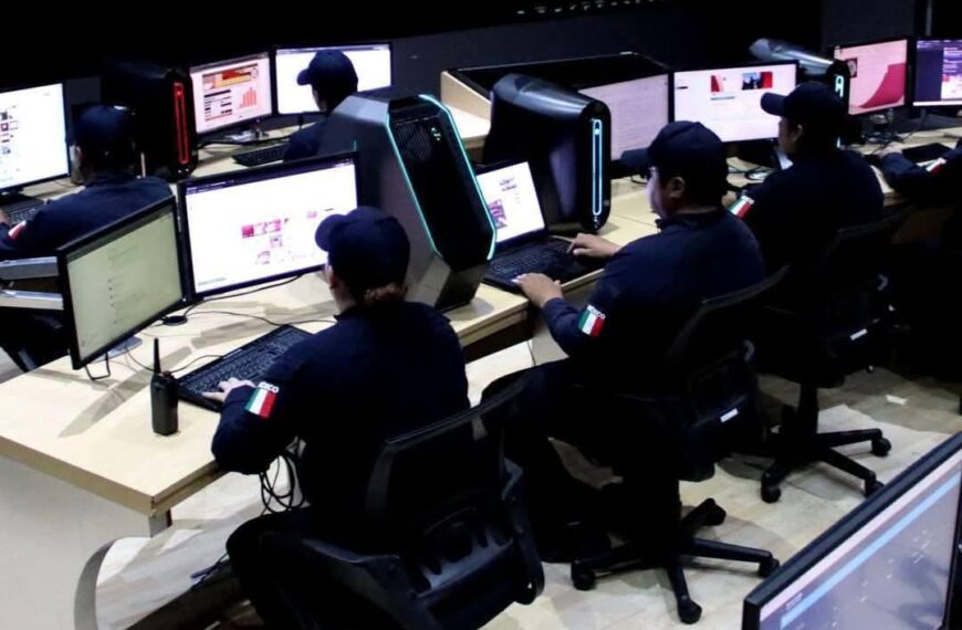 Group of operators in dark uniforms sit at a row of desks with monitors and illuminated PC towers in a computer lab, Italian flag patches on sleeves.