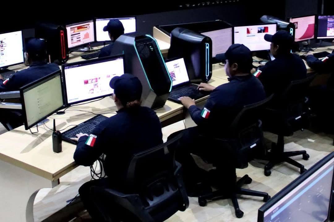 Group of operators in dark uniforms sit at a row of desks with monitors and illuminated PC towers in a computer lab, Italian flag patches on sleeves.