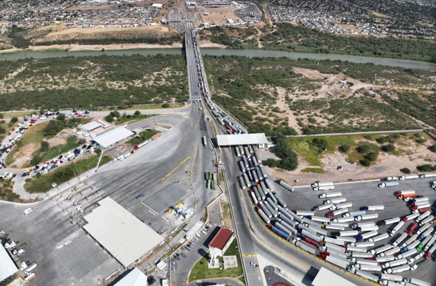 Aerial view of a busy truck checkpoint: long line of semi-trailers on a bridge and approach roads crossing a river into an industrial area.