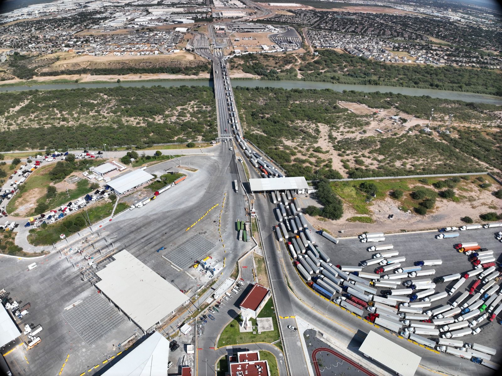 Aerial view of a busy truck checkpoint: long line of semi-trailers on a bridge and approach roads crossing a river into an industrial area.