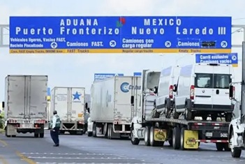 Line of semis waiting at a border crossing under a blue sign reading Aduana Fronterizo Nuevo Laredo.