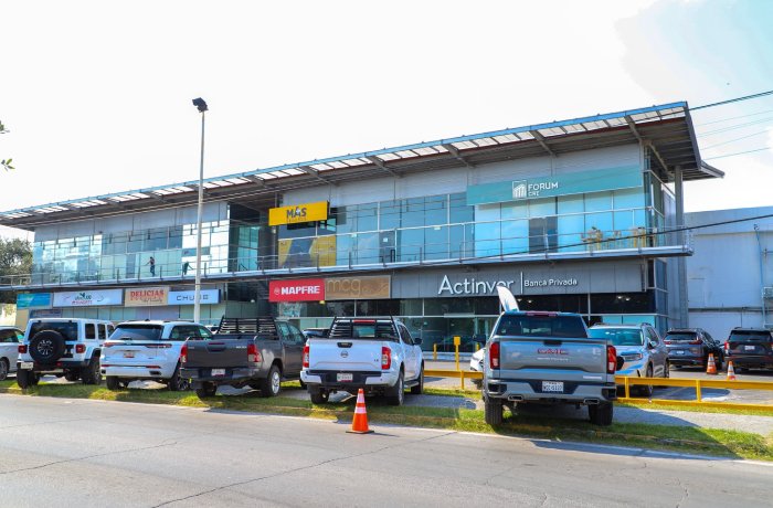 Exterior of a two-story glass-fronted shopping center with multiple storefronts and a parking lot in front. Vehicles parked along the curb.